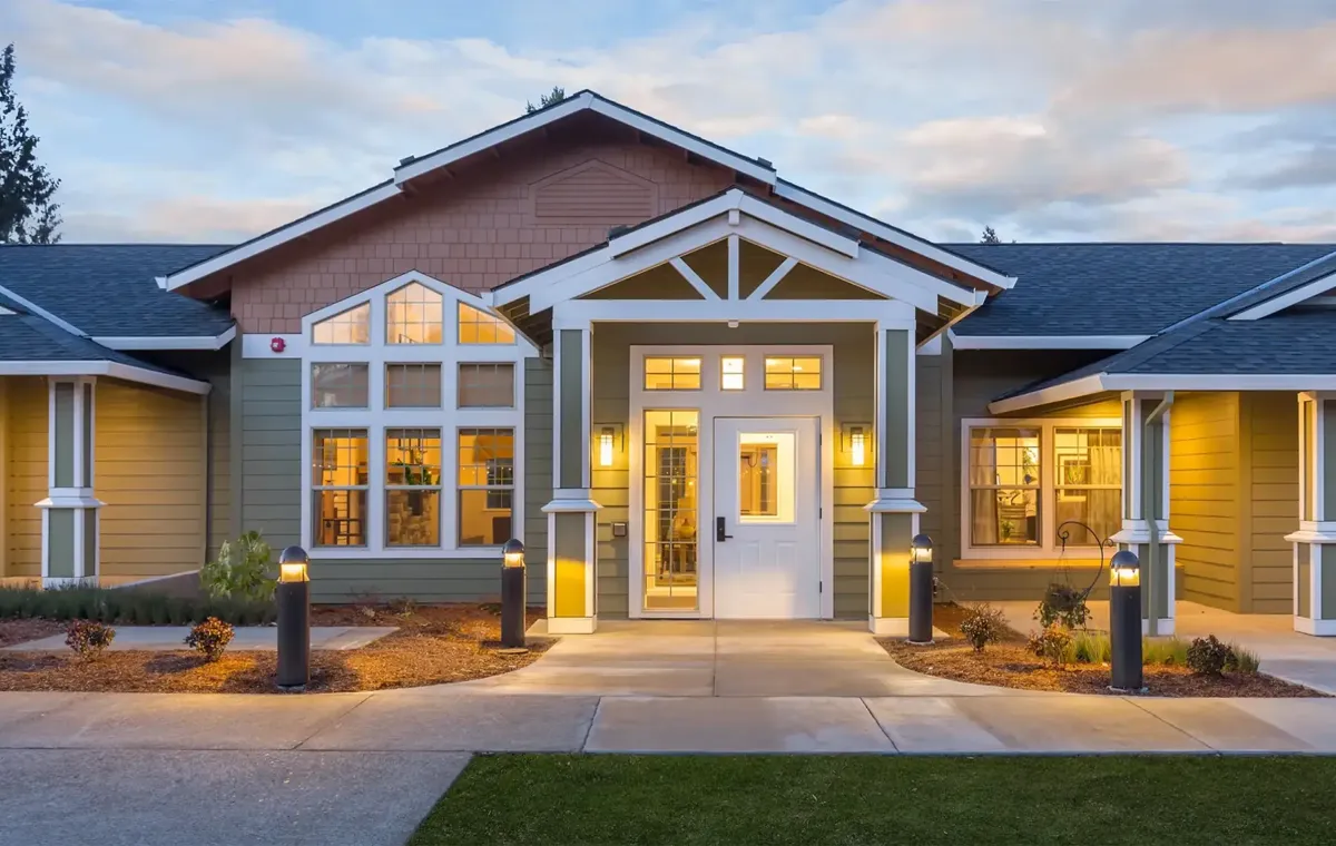A craftsman-style single-story building at dusk, its gabled entry lit by wall sconces and a row of path bollards