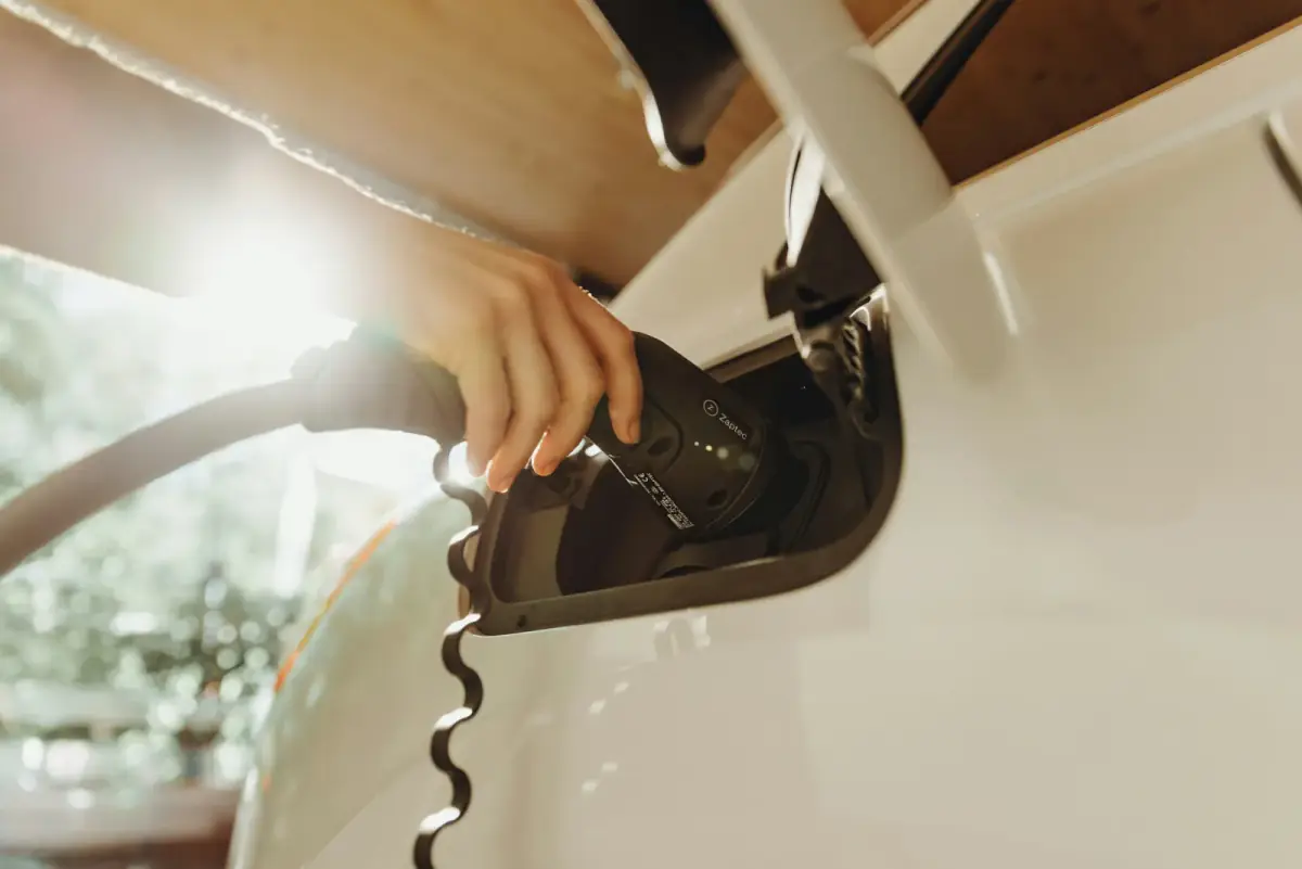 A hand plugging a charging connector into the charge port of a white electric car parked under a timber carport