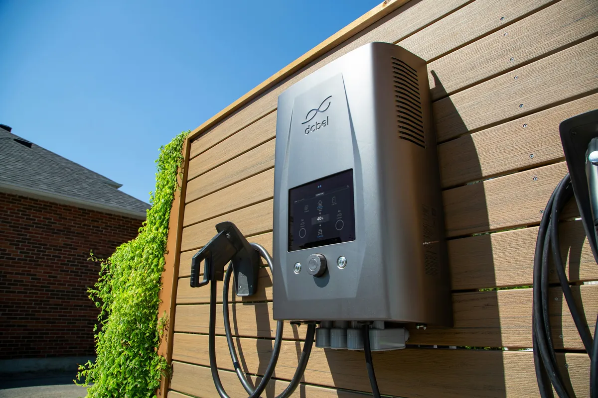 A wall-mounted home EV charger with a touchscreen display on an outdoor slatted fence, its connector holstered beside it