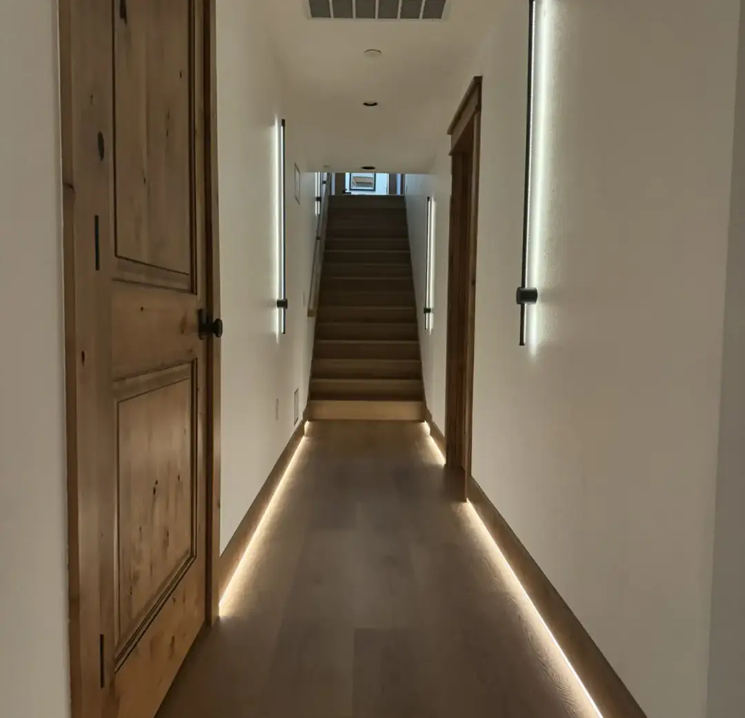 Rewired hallway lit by linear wall sconces, recessed cans, and a continuous LED strip along the baseboard, looking toward the stairs