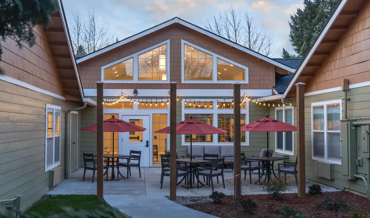 A courtyard at dusk under string lights, with umbrellas and tables and warm light through the gable windows