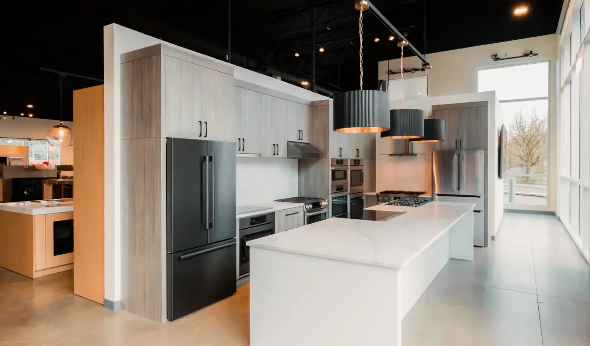 A kitchen appliance showroom under track lighting on an open black ceiling, fluted pendants over a white stone island