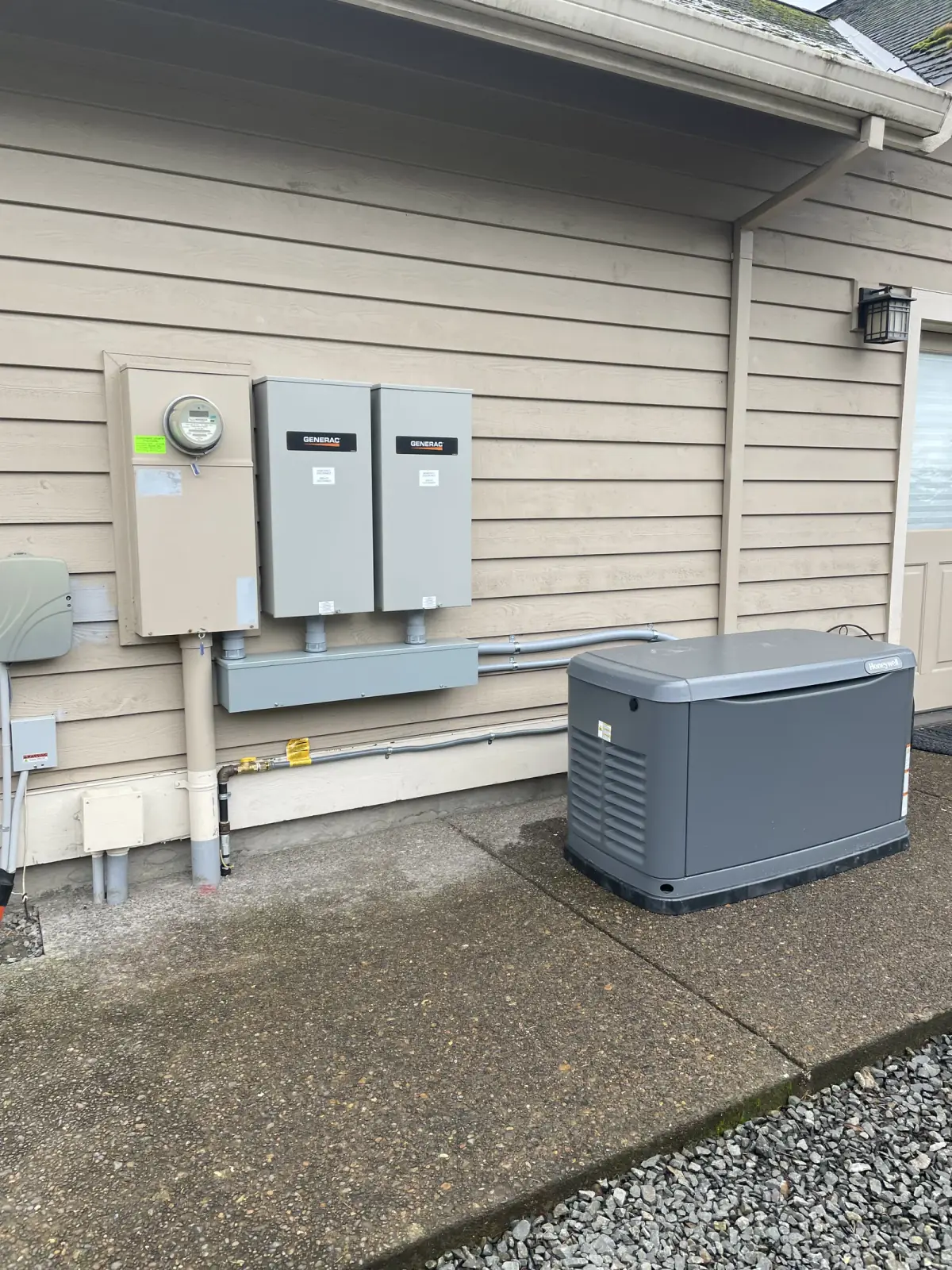 A meter and two transfer switches on a lap-sided wall, with a standby generator on the drive beside them