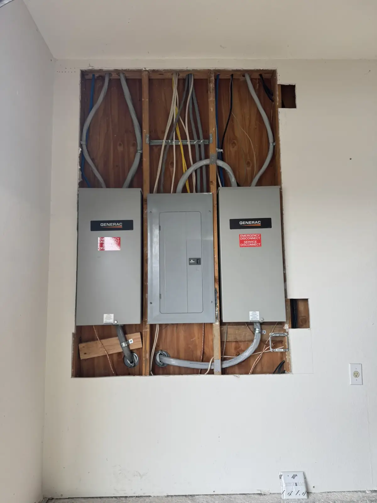 Two Generac transfer switches and a load center in an opened wall, feeders run across the studs