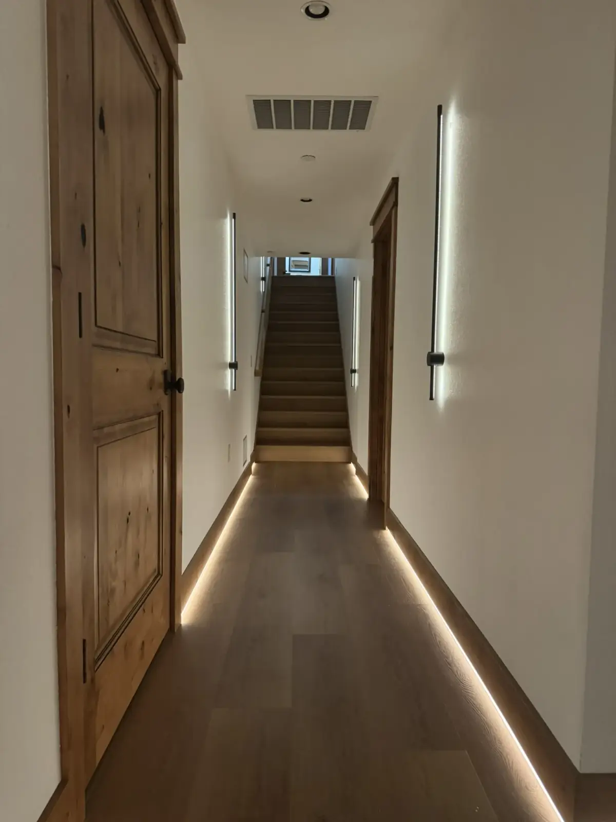 A hallway with linear wall sconces and a continuous LED cove along the floor, leading to a lit stair
