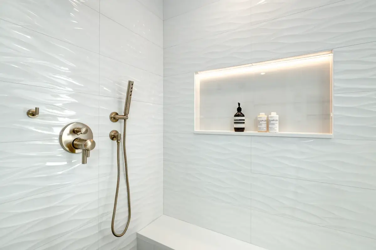 A lit recessed niche in a white tiled shower, beside a wall-mounted mixer and handset