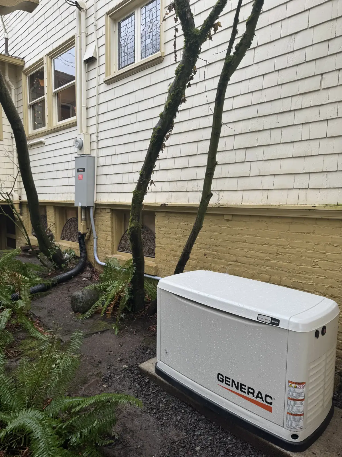 A white standby generator on a pad beside an older shingled house, transfer switch on the wall behind it