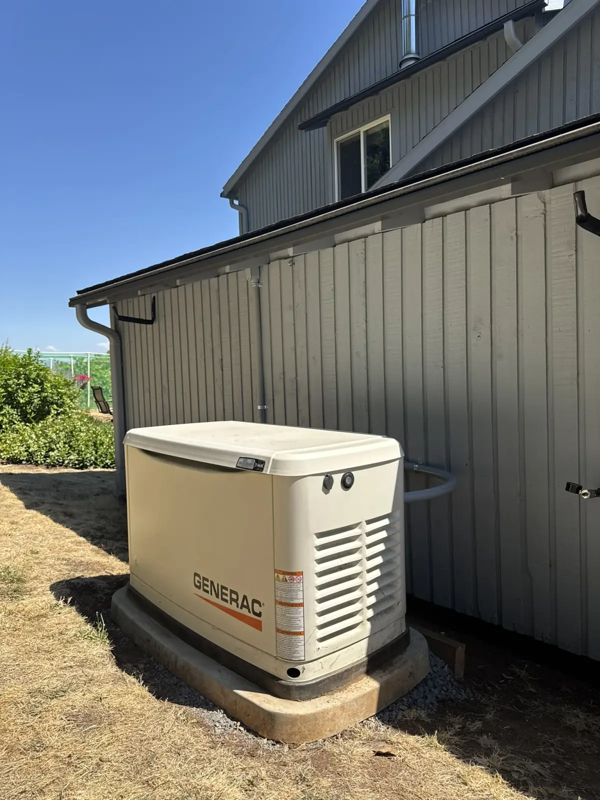 A Generac standby generator on a concrete pad beside a white board-and-batten house