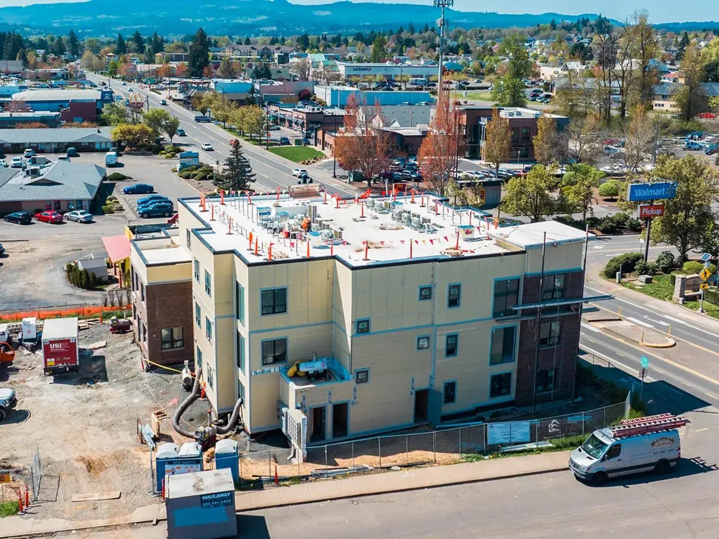 Three-story mixed-use building under construction from the air, rooftop equipment stubbed out and a Multiphase Electric van at the curb