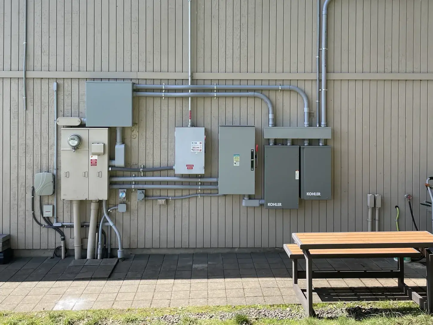 Service equipment across an exterior wall — meter, disconnects, load center and two Kohler transfer switches joined by conduit