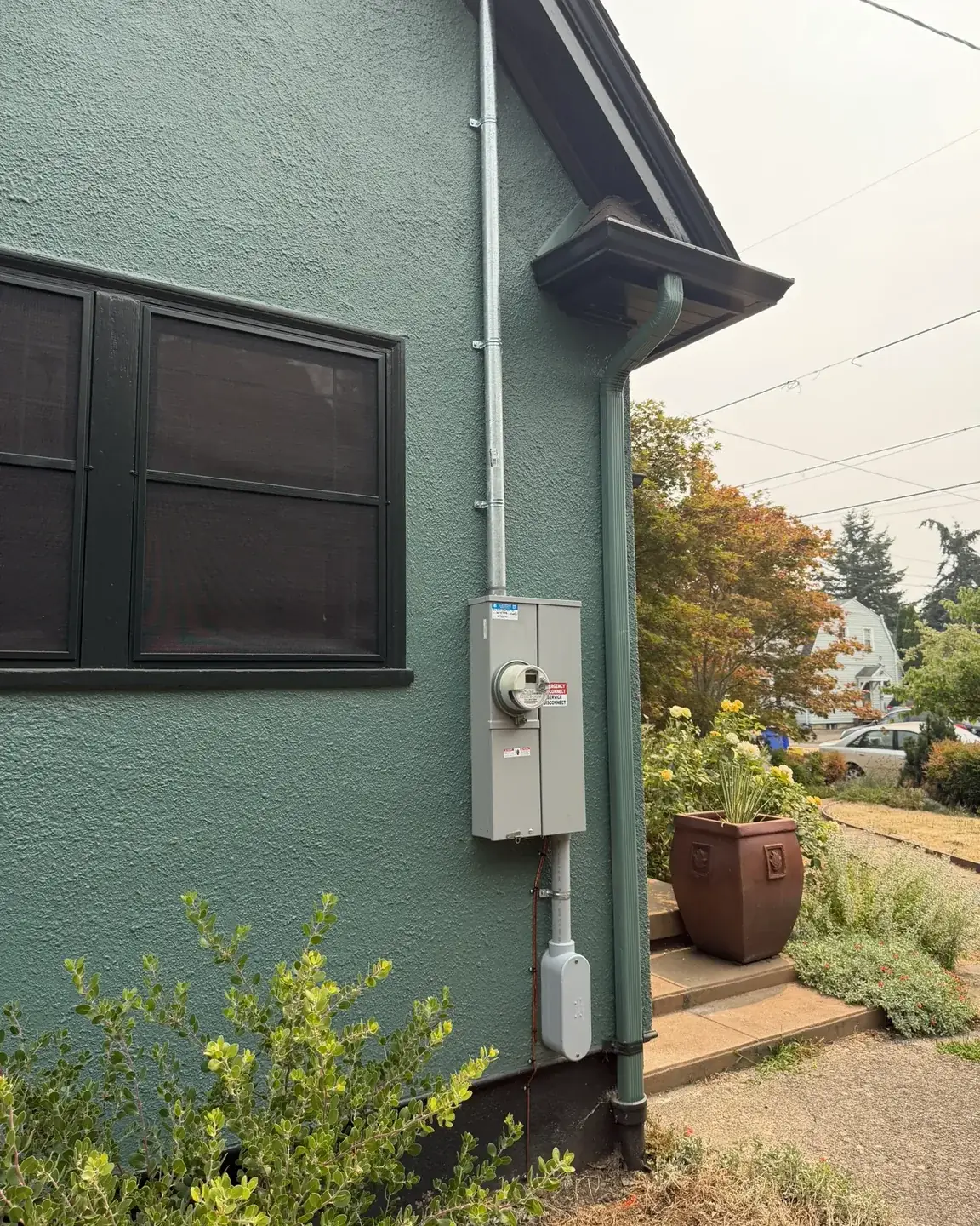 A meter and service disconnect on the teal stucco wall of a house, the service mast run up past the gutter to the roofline