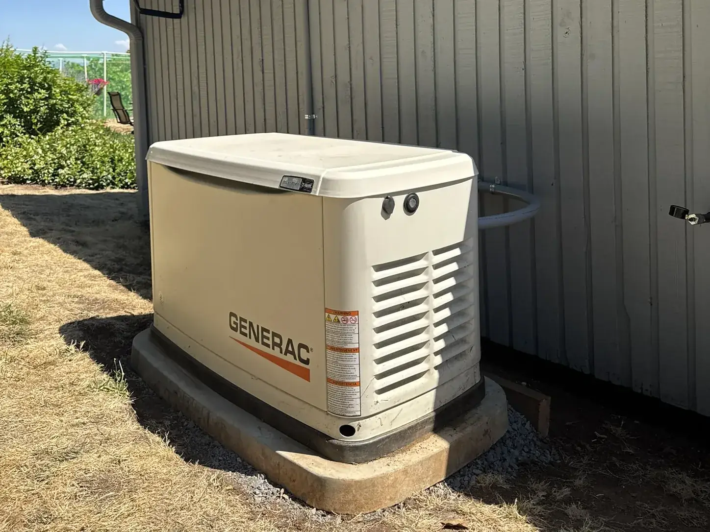 Generac standby generator on a concrete pad beside a board-and-batten farmhouse, fuel line run into the wall behind it