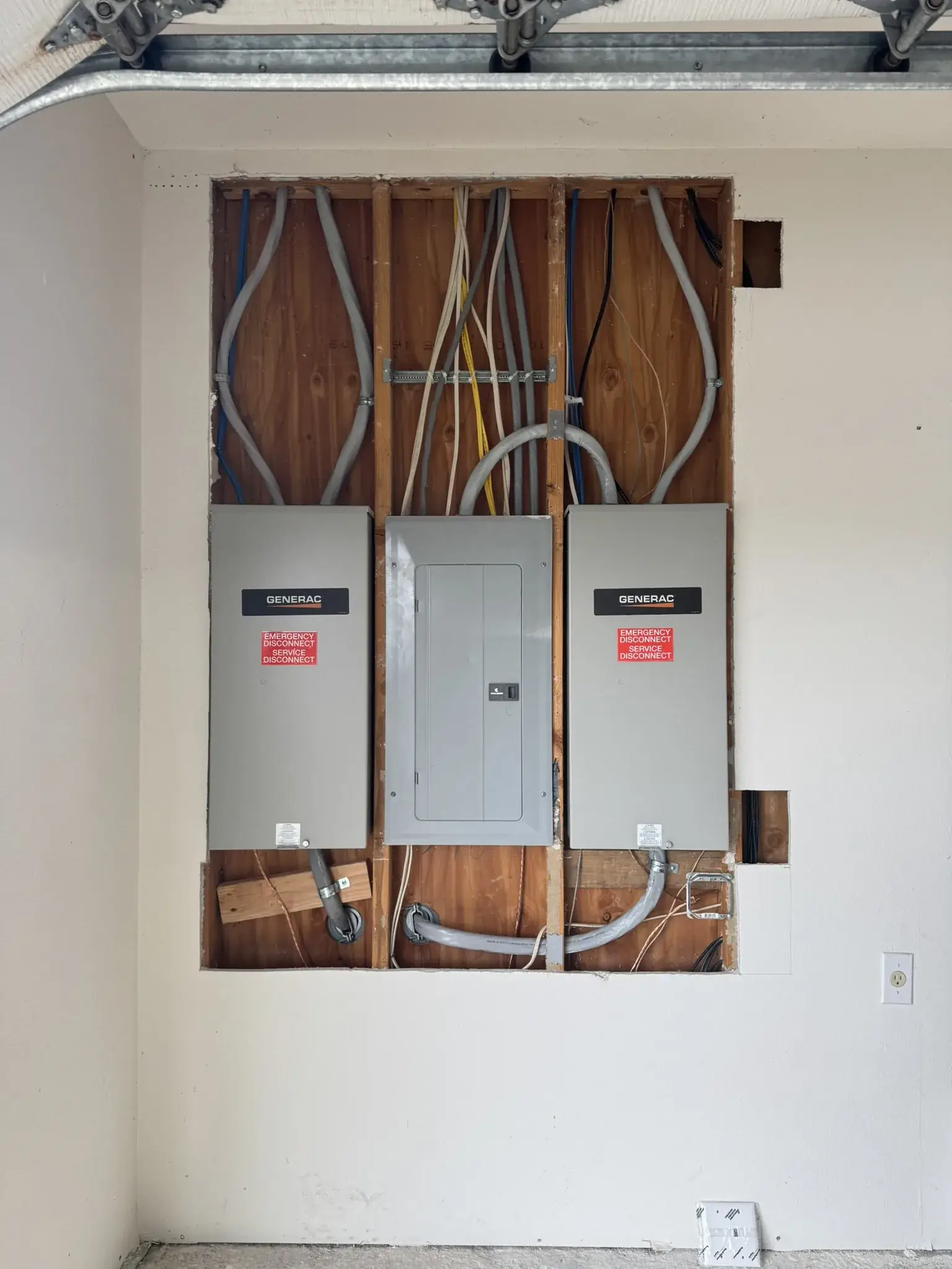Garage wall opened to the studs around a new load center flanked by two Generac transfer switches, feeders and conduit run into the panels